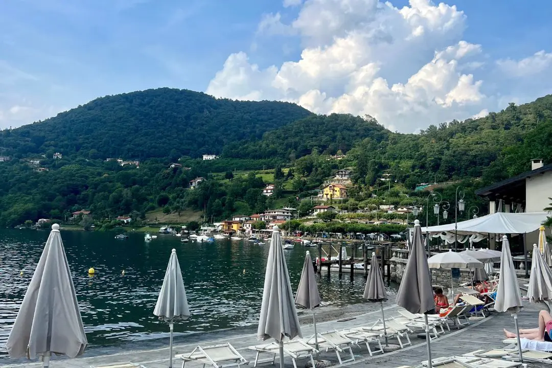 Lakeside view of sun loungers and umbrellas on a beach deck at Lago d’Orta, with green hills and boats in the background